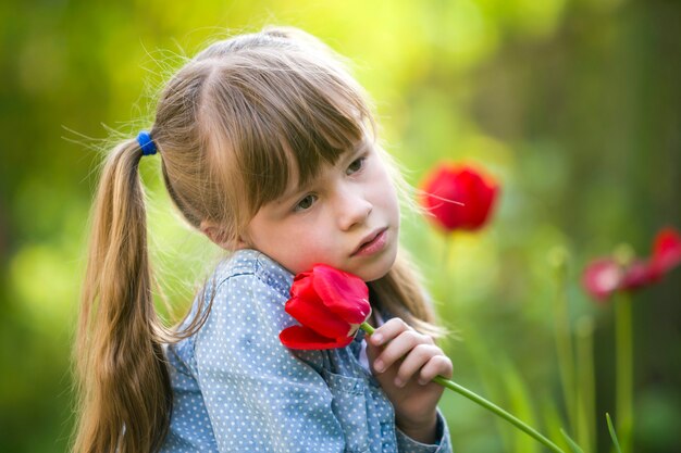 Leuk mooi glimlachend kindmeisje met grijze ogen en lang haar met heldere rode tulpenbloem op vage zonnige groene bokehachtergrond. Love to nature concept.