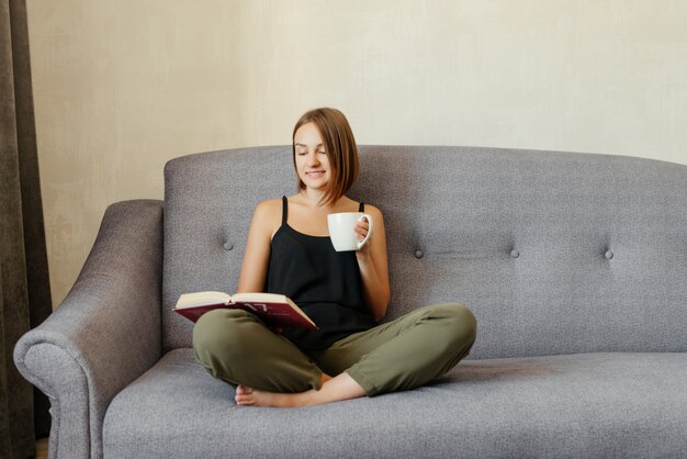 Leuk mooi glimlachend meisje die een boek lezen en thee drinken terwijl thuis het ontspannen op de bank. hobby levensstijl.