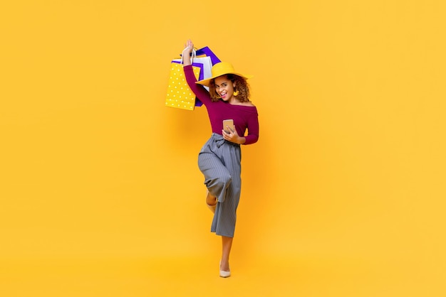 Leuk portret van de volledige lengte van een gelukkige jonge aantrekkelijke shopaholic Afro-Amerikaanse vrouw die boodschappentassen opheft en mobiele telefoon vasthoudt in een geïsoleerde studio gele achtergrond