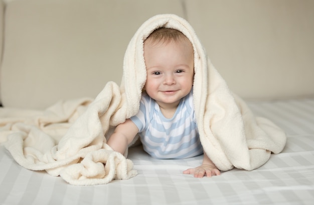 Leuk portret van glimlachende babyjongen die op bed onder witte deken ligt