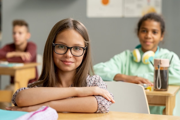 Leuk schoolmeisje met een bril die naar je kijkt bij het bureau