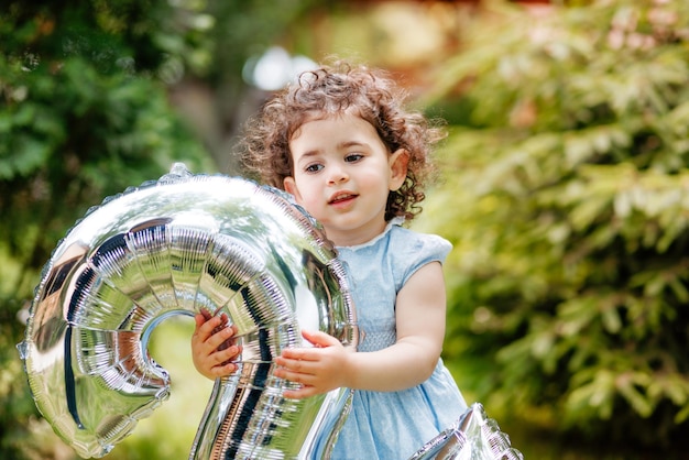 Foto leuk tweejarig meisje met krullend haar dat haar verjaardag viert met een ballonnummer in haar hand.