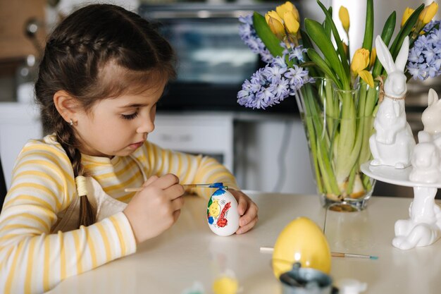 Leuk vierjarig meisje in een beige schort dat op de keukentafel zit en paaseieren schildert.