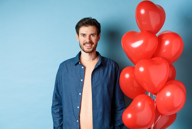 Leuk vriendje op zoek gelukkig en glimlachend, staande in de buurt van Valentijnsdag hart ballonnen op blauwe achtergrond.