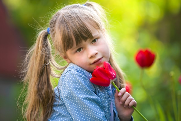 Leuk vrij glimlachend kindmeisje met grijze ogen en lang haar met heldere rode tulpenbloem op vage zonnig groene bokehachtergrond. Graag natuur concept.