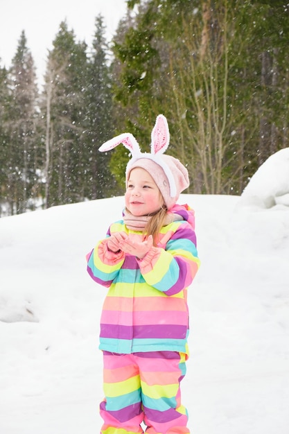 Leuk vrolijk kleutermeisje met caranale konijnenoren die in de sneeuw sneeuw spelen in de winterpret van de paashaas in april