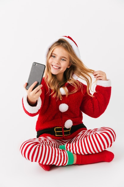 Leuk vrolijk meisje dat geïsoleerde kerstkostuum draagt, mobiele telefoon houdt, een selfie neemt