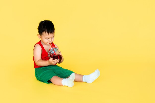 Leuk weinig babyjongen in rode t-shirt en groene kort met kop sap op gele kleurenachtergrond