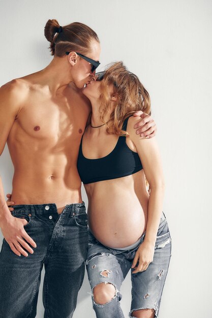 Leuk zwanger koppel zoent in spijkerbroek en zonnebril op een witte achtergrond.