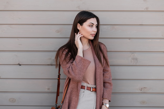 Leuke aantrekkelijke jonge vrouw mannequin in elegante bruine kleding met lederen mode handtas poseren in de buurt van vintage houten gebouw op straat. Vrij fijn meisje in casual outfit buitenshuis. Schoonheidsdame.