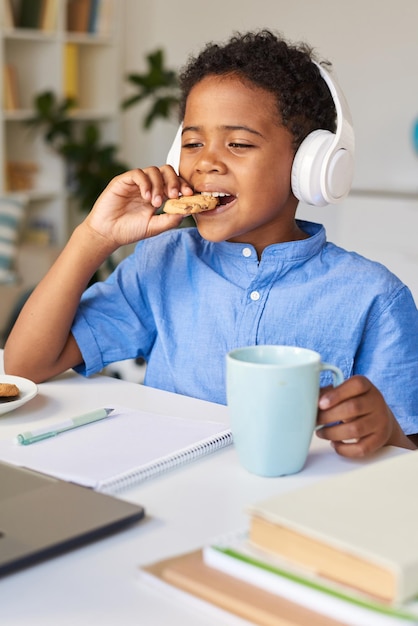 Leuke Afro-Amerikaanse jongen met draadloze koptelefoon die aan tafel zit en koekjes bijt terwijl hij educatieve video's kijkt