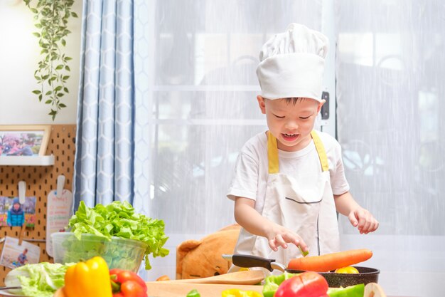 Leuke Aziaat weinig jongenskind chef-kokhoed dragen en schort die pret voorbereidingen treffen, die gezond voedsel in keuken koken