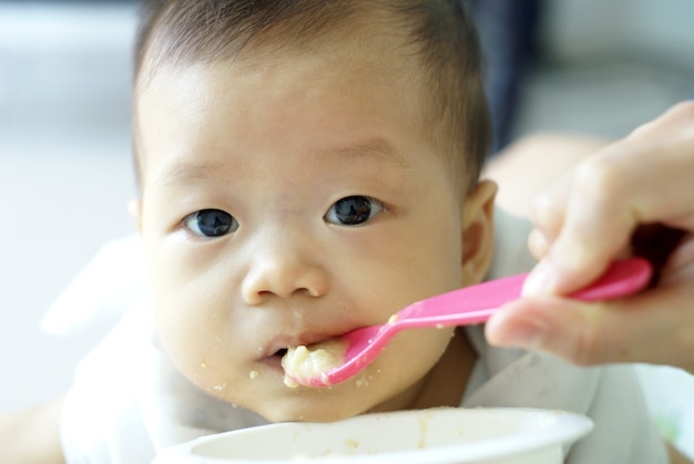 Leuke Aziatische babybaby die wat voedsel met roze lepel in de hand van haar moeder eet.