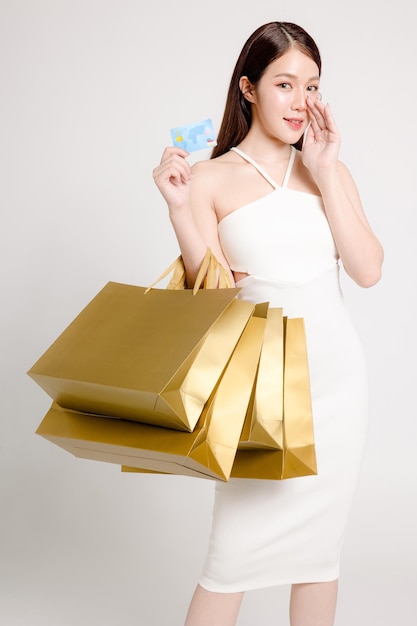 Leuke Aziatische vrouw die glimlacht en gouden boodschappentassen vasthoudt, toont creditcard Mooi vrouwelijk model draagt sexy jurk en natuurlijke make-up op witte achtergrond Cosmetologie wellness zomer verkoop concept