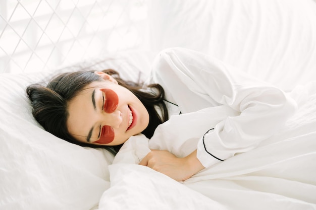 Leuke Aziatische vrouw die pyjama's en collageenogen met deken draagt, ligt thuis op het bed