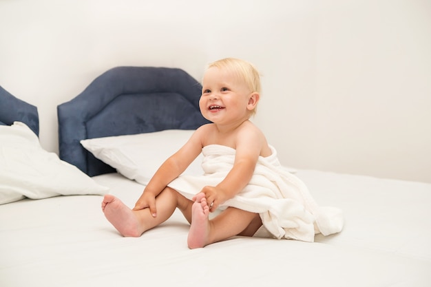 Leuke baby die onder een witte handdoek glimlacht en op het bed zit