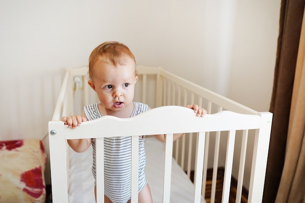 Leuke baby die zich in een wit rond bed bevindt. Witte kinderkamer voor kinderen. Klein meisje leren staan in zijn wieg.