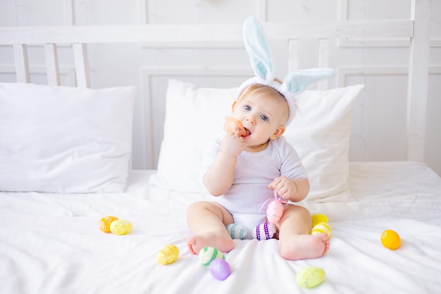 Leuke baby jongen met konijn ooren en kleurrijke eieren op een wit bed thuis spelen of eten eieren kleine blonde baby gelukkig paasconcept