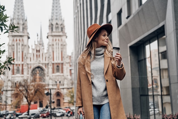 Leuke dag in de stad. Aantrekkelijke jonge vrouw met hoed en jas die een kopje koffie vasthoudt en wegkijkt terwijl ze buiten staat met de kerk op de achtergrond