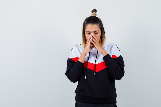 Leuke dame die haar handen op het gezicht houdt, naar beneden kijkt in hoodie en neergeslagen kijkt, vooraanzicht.