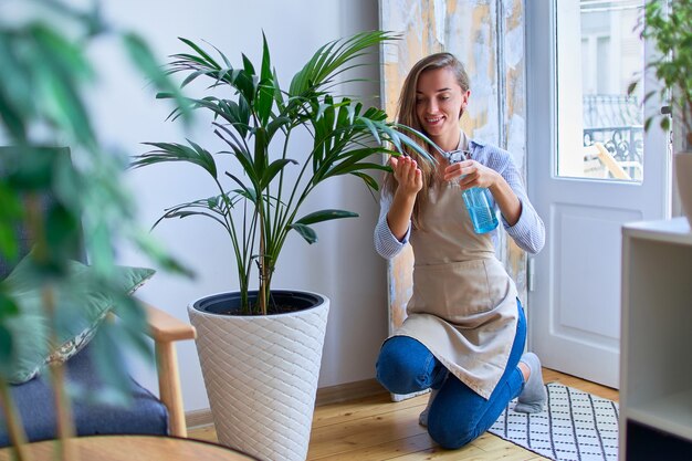 Foto leuke gelukkige jonge glimlachende aantrekkelijke vrouwentuinman in schort die kamerplanten drenken die sproeifles gebruiken