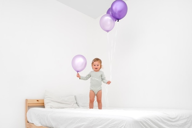 Leuke grappige Kaukasische blonde baby girlpeuter van 12 jaar oud spelen op bed met grote paarse ballonnen van zeer peri trendy kleur op witte achtergrond in moderne Scandinavische interieur thuis