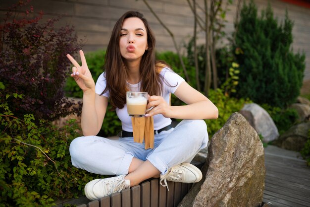 Leuke gratis jonge vrouw geniet van koffie zittend in een mooi zomers park op de steen.