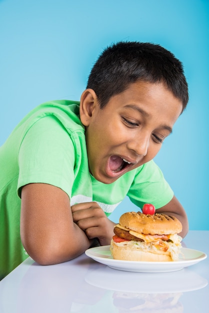 Leuke Indiase jongen die hamburger, kleine Aziatische jongen en hamburger eet, over blauwe achtergrond eating