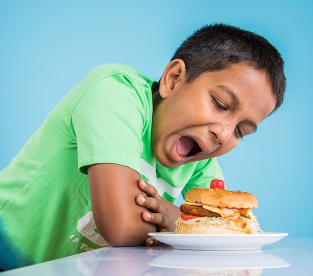 Leuke Indiase jongen die hamburger, kleine Aziatische jongen en hamburger eet, over blauwe achtergrond eating