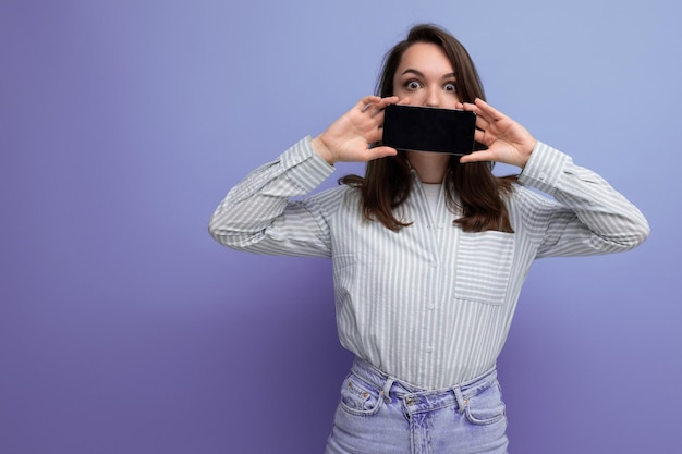 Leuke jonge donkerbruine vrouw in de telefoon van de overhemdsholding met het scherm vooruit op studioachtergrond