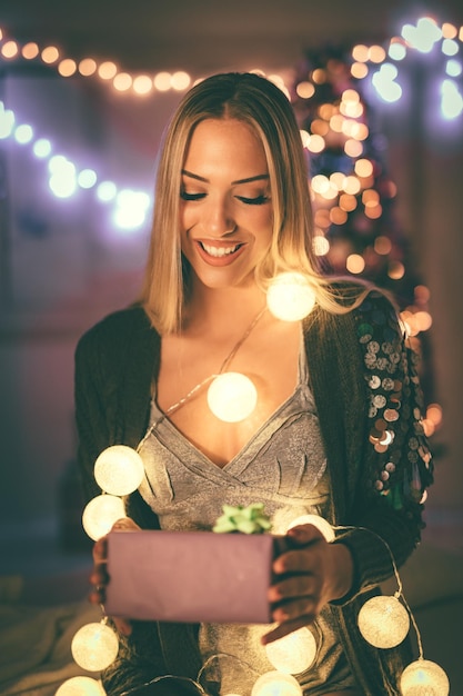 Leuke jonge glimlachende vrouw die een cadeau vasthoudt, omringd met kerstverlichting.