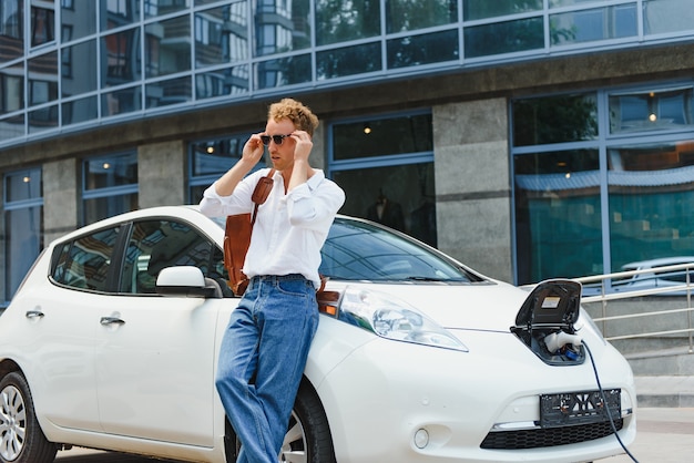 Leuke jonge man in de buurt van een moderne elektrische auto. Het concept van ecologisch vervoer.