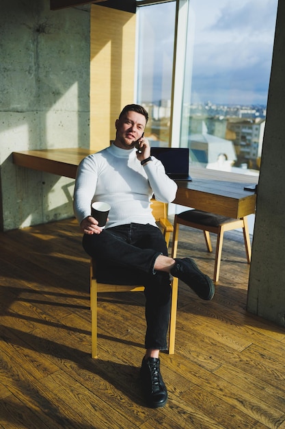 Leuke jonge mannelijke freelancer die koffie drinkt terwijl hij in een lichte werkruimte zit en op afstand werkt Een man in een witte trui en broek werkt vrolijk op kantoor