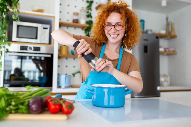 Foto leuke jonge vrouw die kookt en specerijen toevoegt aan de maaltijd lacht en tijd doorbrengt in de keuken