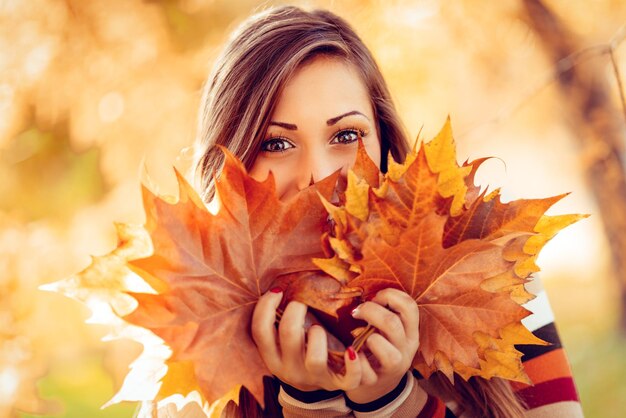 Leuke jonge vrouw genieten in zonnig bos in herfstkleuren. Ze houdt veel bladeren vast en kijkt naar de camera achter bladeren.