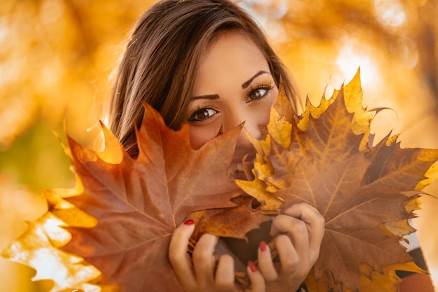 Leuke jonge vrouw genieten in zonnig bos in herfstkleuren. Ze houdt veel gevallen bladeren vast en kijkt naar de camera erachter.