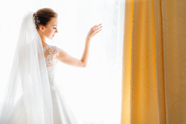 Leuke jonge vrouw in een witte jurk bij het raam van het huis Ochtend van de bruid op de trouwdag Gelukkig meisje wacht op een ontmoeting met de bruidegom Achteraanzicht