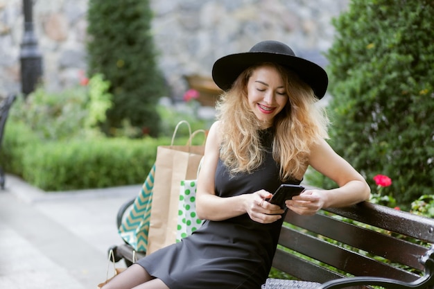 Leuke jonge vrouw in een zwarte vilten hoed en jurk met boodschappentas zit op een bankje in het stadspark en gebruikt smartphone
