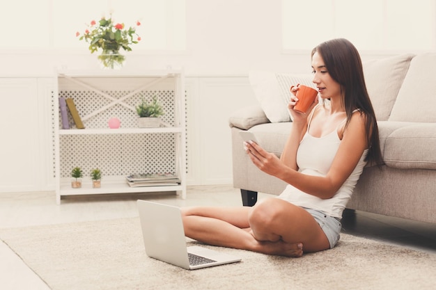 Leuke jonge vrouw messaging op mobiel. Meisje zittend op de vloer met laptop en koffie drinken, kopieer ruimte