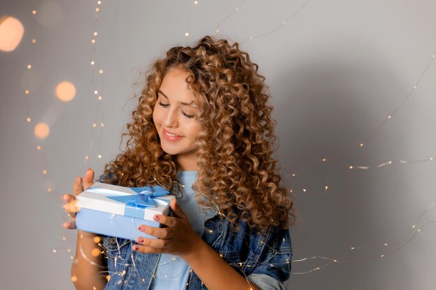 Leuke jonge vrouw met blauwe ogen en blond krullend haar in denim kleding houdt een geschenkdoos in haar handen. meisje opent het geschenk. effen grijze achtergrond. ruimte voor tekst. Hoge kwaliteit foto