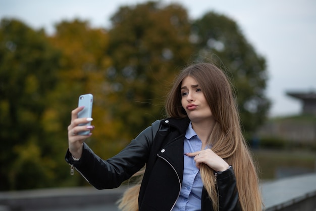 Leuke jonge vrouw met lang haar die grimassen trekt en zelfportret maakt op mobiele telefoon in het park
