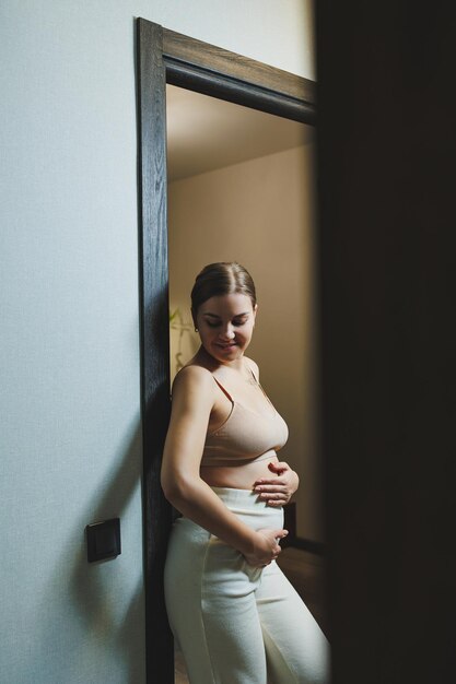 Leuke jonge zwangere vrouw in een top en casual zwangerschapskleding Leuke zwangere vrouw die haar buik thuis knuffelt