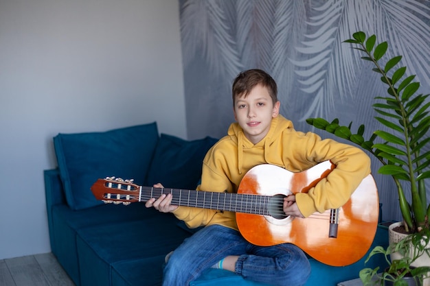 Leuke jongen die akoestische gitaar speelt, gekleed in een casual jeans en gele sweater op de gezellige bank thuis