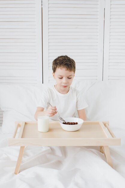 Leuke jongen die de ballen van de ontbijtchocolade zit die in bed met wit linnen zit