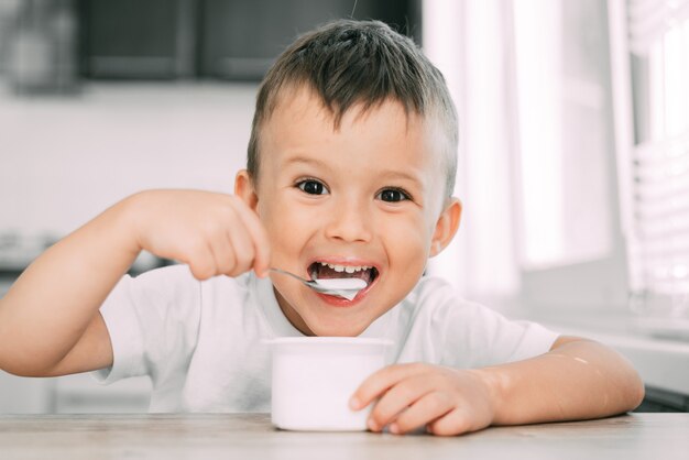 Leuke jongen in de keuken die yoghurt eet in lichte kleren uit een witte yoghurtcontainer, een plek voor reclame