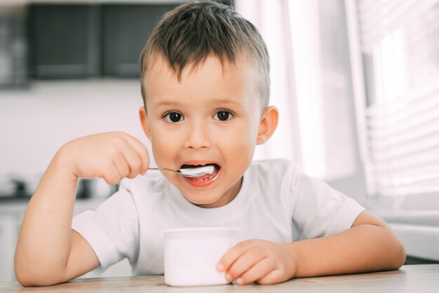 Leuke jongen in de keuken die yoghurt eet in lichte kleren uit een witte yoghurtcontainer, een plek voor reclame