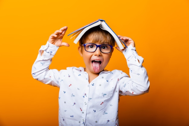 Leuke jongen in glazen met een boek op zijn hoofd toont tong op gele achtergrond