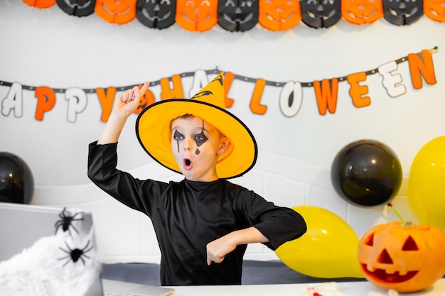 Foto leuke jongen in kostuum zit achter tafel in halloween-thema ingerichte kamer halloween van covid19