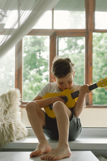 Leuke jongen leert de gele Ukelele-gitaar te spelen op de vensterbank bij het raam Gezellig huis Zomervakantie levensstijl