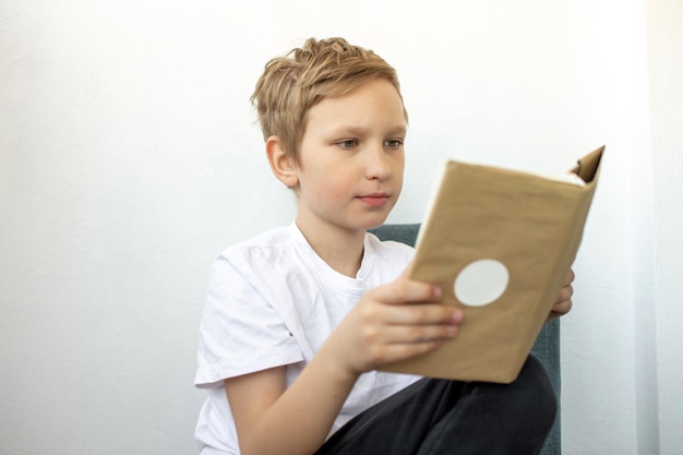 Leuke jongen leest zorgvuldig een boek terwijl hij op een stoel in de kamer zit